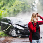 A young woman with smartphone by the damaged car after a car accident, making a phone call.