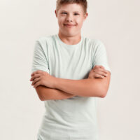 Teenaged disabled boy with Down syndrome smiling at camera while posing, standing with arms crossed isolated over white background